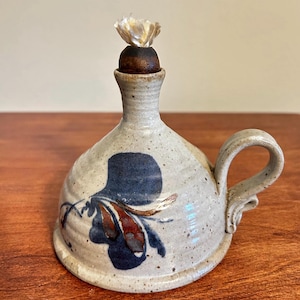 May include: Handmade ceramic oil lamp with a speckled, off-white glaze. The lamp features a dark blue floral design and a small handle. A wooden stopper with a white flower sits on top. The lamp is approximately 5 inches tall.