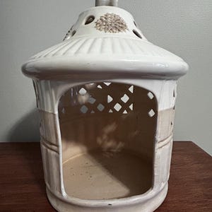May include: Cream-colored ceramic lantern with a domed top and open sides. The lantern features decorative cutouts and a handle at the top. A floral design is embossed on the top. The lantern is designed to hold a candle.