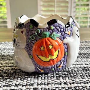 May include: A ceramic bowl featuring a raised orange jack-o'-lantern with star-shaped eyes, a white cat, and a white ghost against a purple brick-patterned background. The rim has a scalloped edge with black and white accents.