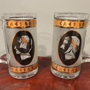 May include: Two clear glass beer mugs featuring a frosted finish and a gold band at the top and bottom with the text CAESARS. Each mug displays a central black oval containing a white profile illustration of a Roman figure wearing a laurel wreath.