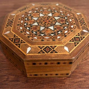 May include: An octagonal wooden box featuring intricate marquetry with geometric patterns in various shades of brown, black, and tan wood. The lid is decorated with a central star motif inlaid with white mother-of-pearl accents throughout.