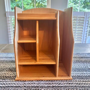May include: A light-colored wooden desktop organizer featuring a vertical divider, a top shelf, and two lower shelves. The structure is constructed from smooth, natural-toned wood with visible grain patterns, providing multiple open compartments for storage.