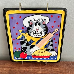 Può includere: Un piatto quadrato in ceramica raffigurante un gatto tigrato grigio e bianco con un cappello da chef bianco e una sciarpa gialla. Il gatto tiene un mattarello di legno sopra una torta gialla. Lo sfondo è viola con pois rossi, bordo giallo e nero.