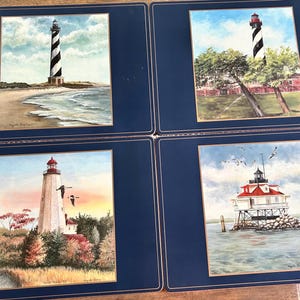 May include: A set of four rectangular placemats with dark blue borders and gold trim. Each placemat features a different lighthouse painting: one on a beach, one near trees, one at sunset with birds, and one offshore on a rocky foundation.