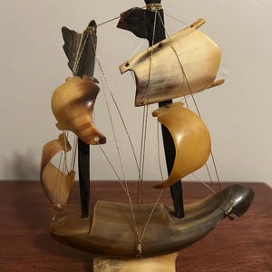 Könnte beinhalten: Eine handgefertigte Miniatur-Segelboot-Skulptur. Das Boot hat einen dunkelbraunen Rumpf, Masten und Segel aus hellbraunem Material. Goldfarbene Schnur wird für das Takelwerk verwendet, und die Skulptur steht auf einem rechteckigen Sockel.