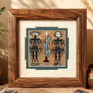 May include: A square framed sand art piece featuring two stylized figures flanking a central blue plant-like form. The artwork is set against a tan background, enclosed in a dark teal mat with notched edges, and encased in a rustic brown wood frame.