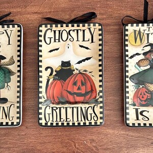 May include: Three rectangular ceramic plaques with black ribbon hangers. The first reads Happy Haunting with a pumpkin-headed figure. The second reads Ghostly Greetings with a black cat and pumpkins. The third reads The Witch Is In with a witch figure.