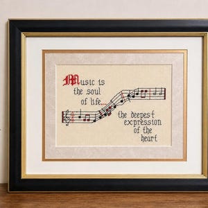 Może przedstawiać: Oprawiony haft krzyżykowy na beżowej tkaninie z czarno-czerwonym napisem Music is the soul of life... the deepest expression of the heart, któremu towarzyszy czarno-czerwona pięciolinia z nutami w ozdobnej ramie.