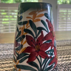 以下が含まれることがあります： 白いベースに手描きの花柄が施された円錐形の陶器製花瓶。デザインには、大きな赤いユリのような花、濃い緑色の葉、細い黄色の小枝が含まれています。花瓶の縁は濃い緑色の帯で仕上げられています。
