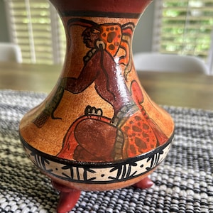 May include: A terracotta-coloured ceramic vase featuring a hand-painted figure in profile wearing an ornate headdress. The vessel has a flared rim, a rounded body, and three short, sturdy feet. A decorative band with black geometric patterns encircles the base.