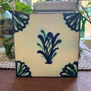 Peut inclure: Un carreau de céramique carré avec un fond crème présentant un motif floral bleu foncé et vert peint à la main au centre. Les quatre coins du carreau sont décorés de motifs en forme d'éventail assortis en bleu foncé et vert.
