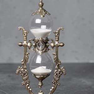 May include: An antique-style hourglass with a bronze-colored metal frame. The clear glass bulbs contain white sand. The hourglass is supported by an ornate base and decorative side supports. A classic decorative item.