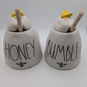 May include: Two white ceramic honey pots with lids, each featuring a yellow bee topper and a wooden honey dipper. The left pot is labeled HONEY and the right pot is labeled BUMBLE in black lettering, with a small black bee graphic below each word.
