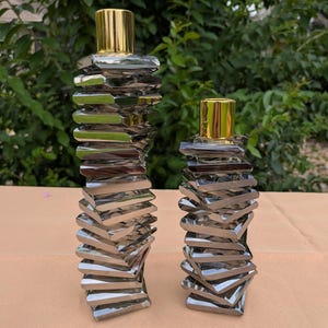 May include: Two decorative candle holders featuring a stacked, geometric design made of reflective silver-toned square plates. Each holder is topped with a smooth, cylindrical gold-toned metal cap, creating a tiered, spiraling appearance.