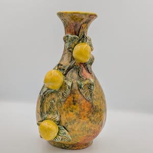 May include: A ceramic vase with a textured, mottled finish in shades of yellow, orange, and brown. The vase is adorned with three-dimensional yellow fruit and green leaves, adding a naturalistic touch to the decorative piece.