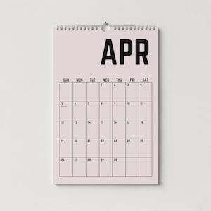 Peut inclure: Un calendrier mural rose clair pour le mois d'avril, avec du texte noir. Le mot "APR" est en haut, avec une grille de dates en dessous. Le calendrier est maintenu par une reliure en spirale argent&eacute;e et un petit trou pour l'accrocher.