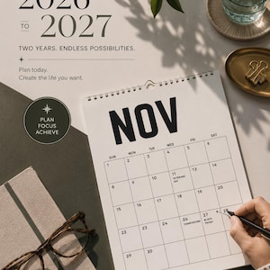 Peut inclure: Un calendrier ouvert sur le mois de novembre, avec les ann&eacute;es 2026-2027 affich&eacute;es en haut. Le texte &laquo; Planifiez aujourd'hui. Cr&eacute;ez la vie que vous voulez. &raquo; est visible. Une personne &eacute;crit sur le calendrier avec un stylo.