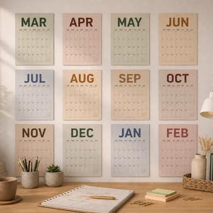 May include: A collection of twelve monthly calendars, each with a different pastel background color and month name. The calendars are displayed on a neutral-colored wall above a wooden desk. A desk lamp, stationery, and a coffee cup are also visible.