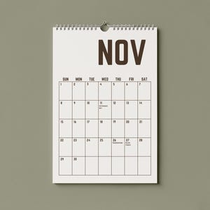 May include: A white spiral-bound calendar for November hangs against a muted green background. The word "NOV" is printed in bold brown letters at the top. The calendar grid displays the days of the month, with dates and holidays.