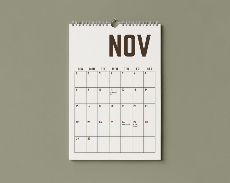 May include: A white spiral-bound calendar for November hangs against a muted green background. The word "NOV" is printed in bold brown letters at the top. The calendar grid displays the days of the month, with dates and holidays.