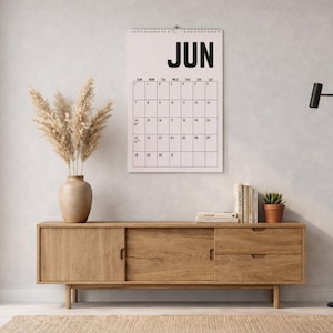 Pode incluir: Cena interior minimalista com um aparador de madeira, um calend&aacute;rio exibindo "JUN" em letras pretas grandes e um candeeiro de p&eacute; preto. Um vaso com capim-dos-pampas seco e uma pequena planta em vaso completam a decora&ccedil;&atilde;o.