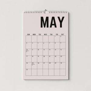 Peut inclure: Calendrier mural de mai avec un fond rose clair et du texte noir. Le mot "MAI" est en haut, avec les jours de la semaine en dessous. La grille du calendrier est visible, avec les dates et les jours f&eacute;ri&eacute;s marqu&eacute;s.
