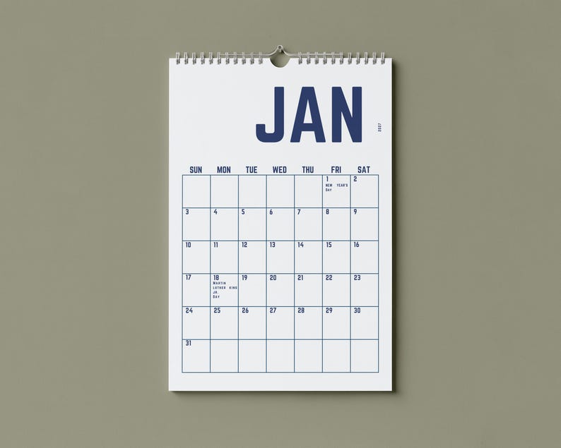 May include: A white wall calendar for January 2017, hanging against a muted green background. The calendar features a grid layout with the days of the week across the top and dates in blue. The word "JAN" is prominently displayed at the top.