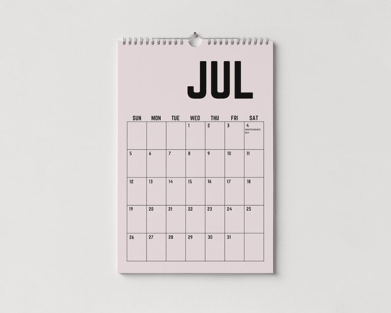 Peut inclure: Calendrier mural de juillet, reli&eacute; par une spirale en haut. Le mot "JUL" est en noir gras. La grille du calendrier affiche les jours de la semaine et les dates, avec le 4 marqu&eacute; comme Jour de l'Ind&eacute;pendance.