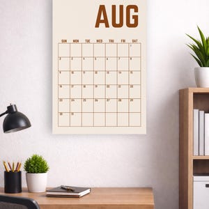 May include: A wall calendar for August, with a cream-colored background and brown grid lines. The word "AUG" is at the top. The calendar is hanging above a wooden desk with a black desk lamp and a small potted plant.
