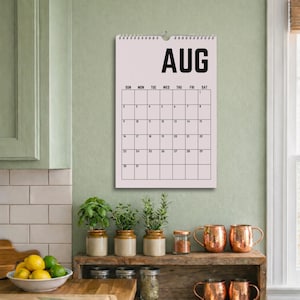 Peut inclure: Un calendrier d'ao&ucirc;t rose clair est accroch&eacute; au mur au-dessus d'une &eacute;tag&egrave;re en bois avec des mugs en cuivre et des plantes en pot. Le calendrier affiche les jours du mois en texte noir. Le mot "AUG" est en haut du calendrier.