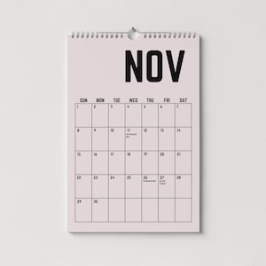 Peut inclure: Un calendrier mural rose pour le mois de novembre avec une reliure &agrave; spirale en haut. Le mot "NOV" est imprim&eacute; en lettres noires en gras. La grille du calendrier affiche les jours du mois, avec les dates et du texte.