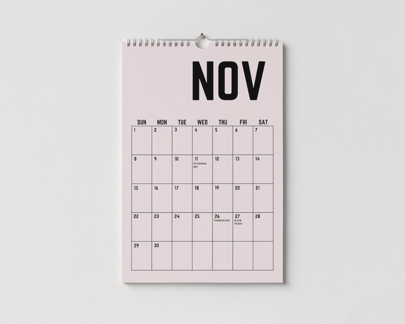 Peut inclure: Un calendrier mural rose pour le mois de novembre avec une reliure &agrave; spirale en haut. Le mot "NOV" est imprim&eacute; en lettres noires en gras. La grille du calendrier affiche les jours du mois, avec les dates et du texte.