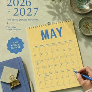 Pode incluir: Um calend&aacute;rio amarelo para maio de 2026-2027 com texto azul, um caderno azul e um clip dourado. O calend&aacute;rio tem encaderna&ccedil;&atilde;o em espiral e uma pessoa segura uma caneta azul. O texto inclui "Plan Focus Achieve".