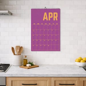 Pode incluir: Um calend&aacute;rio de parede roxo para abril est&aacute; pendurado em uma cozinha. O calend&aacute;rio tem um layout de grade com letras douradas. Os elementos da cozinha incluem um fog&atilde;o, uma panela, utens&iacute;lios e uma tigela de lim&otilde;es.
