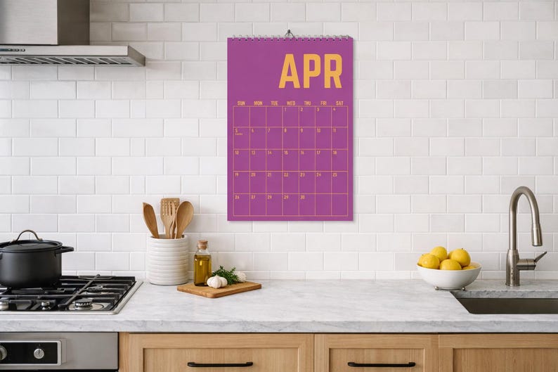 Pode incluir: Um calend&aacute;rio de parede roxo para abril est&aacute; pendurado em uma cozinha. O calend&aacute;rio tem um layout de grade com letras douradas. Os elementos da cozinha incluem um fog&atilde;o, uma panela, utens&iacute;lios e uma tigela de lim&otilde;es.