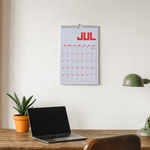 Pode incluir: Calend&aacute;rio de parede de julho com texto e grade vermelhos. Um laptop, uma lumin&aacute;ria de mesa verde e uma planta em vaso est&atilde;o em uma mesa de madeira. O fundo &eacute; uma parede branca com uma coluna de tijolos.