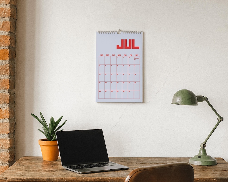 Pode incluir: Calend&aacute;rio de parede de julho com texto e grade vermelhos. Um laptop, uma lumin&aacute;ria de mesa verde e uma planta em vaso est&atilde;o em uma mesa de madeira. O fundo &eacute; uma parede branca com uma coluna de tijolos.