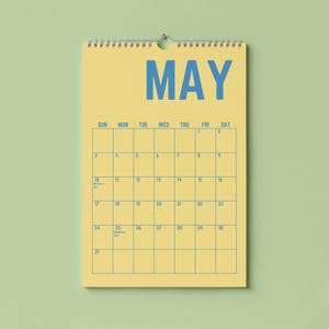 Pode incluir: Um calend&aacute;rio de parede amarelo para maio, com texto e linhas de grade azuis. A palavra "MAY" &eacute; exibida de forma proeminente no topo. O calend&aacute;rio est&aacute; pendurado em um fundo verde claro.