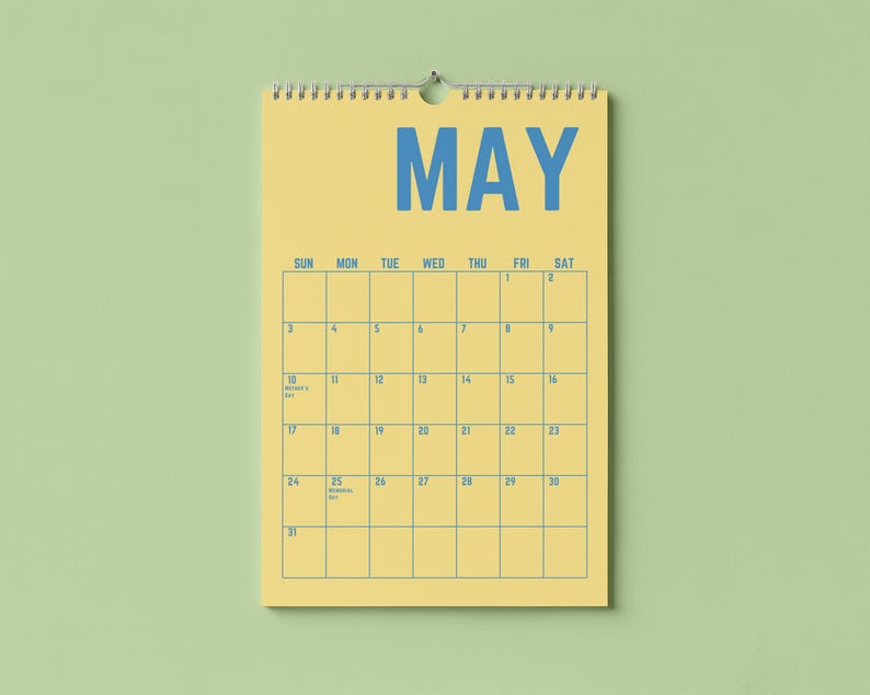 Pode incluir: Um calend&aacute;rio de parede amarelo para maio, com texto e linhas de grade azuis. A palavra "MAY" &eacute; exibida de forma proeminente no topo. O calend&aacute;rio est&aacute; pendurado em um fundo verde claro.