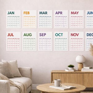 Pu&ograve; includere: Un set di dodici stampe di calendario da parete disposte su due file, ognuna con il nome del mese in un colore distinto e una griglia dei giorni. I mesi da JAN a DEC sono visualizzati con testo colorato e linee di griglia nere su carta.