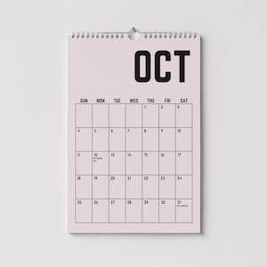 Peut inclure: Un calendrier mural rose pour le mois d'octobre, avec une reliure &agrave; spirale en haut. Le mot "OCT" est imprim&eacute; en lettres noires en gras. La grille du calendrier affiche les jours du mois, avec les dates et quelques jours sp&eacute;ciaux.