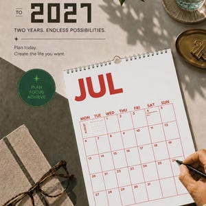 Pu&ograve; includere: Un calendario da parete per luglio che mostra una griglia con testo e linee rosse. Il testo recita 2026 TO 2027, TWO YEARS. ENDLESS POSSIBILITIES. Plan today. Create the life you want. PLAN FOCUS ACHIEVE. Una mano tiene una penna nera.