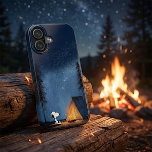 May include: A dark blue phone case featuring a camping scene with Snoopy. The case depicts a tent, campfire, and a starry night sky. The phone case is resting on a wooden log, with a campfire burning in the background.