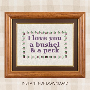 Peut inclure: Broderie au point de croix encadrée. Le texte "I love you a bushel & a peck" est brodé en violet, entouré d'une bordure de points verts et roses. Le cadre a une bordure intérieure marron et un cadre extérieur marron clair.