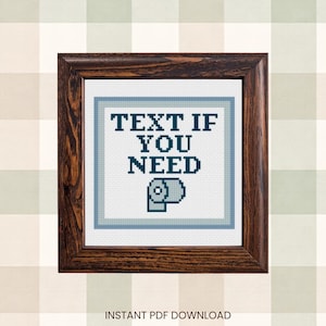 May include: A framed cross-stitch artwork with a dark wood frame. The artwork features the text "TEXT IF YOU NEED" above a toilet paper roll icon, all in dark blue on a light blue and white background. The background is a light beige and green plaid.
