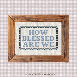 May include: A framed cross-stitch piece with the words "HOW BLESSED ARE WE" in light blue. The text is framed by a dark blue and black border, set against a white background. The frame is made of rustic, light brown wood.