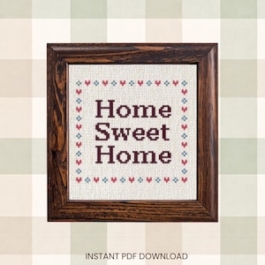 Pode incluir: Um ponto de cruz emoldurado com as palavras "Home Sweet Home" em roxo escuro. A moldura é de madeira escura. O ponto de cruz tem uma borda de pequenos corações e diamantes vermelhos e azuis. O fundo é um padrão xadrez.