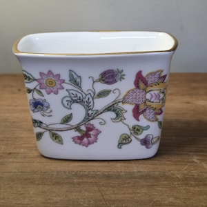 May include: A white ceramic square vase with a gold rim, decorated with a floral pattern in pink, purple, and blue. The design features stylized flowers and leaves, creating a vintage aesthetic.