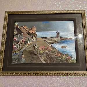 May include: A framed landscape print featuring a coastal village scene with stone buildings, a harbor wall, and a small boat on the water. The artwork is enclosed in a rectangular gold-toned frame with a dark brown matting border surrounding the image.