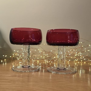 Op de afbeelding: Twee bijpassende coupes met diep magentakleurige kelken en helderglazen stelen en ronde voeten. De kelken hebben een brede, ronde vorm met een gladde rand, terwijl de stelen recht en cilindervormig zijn en op platte voeten staan.